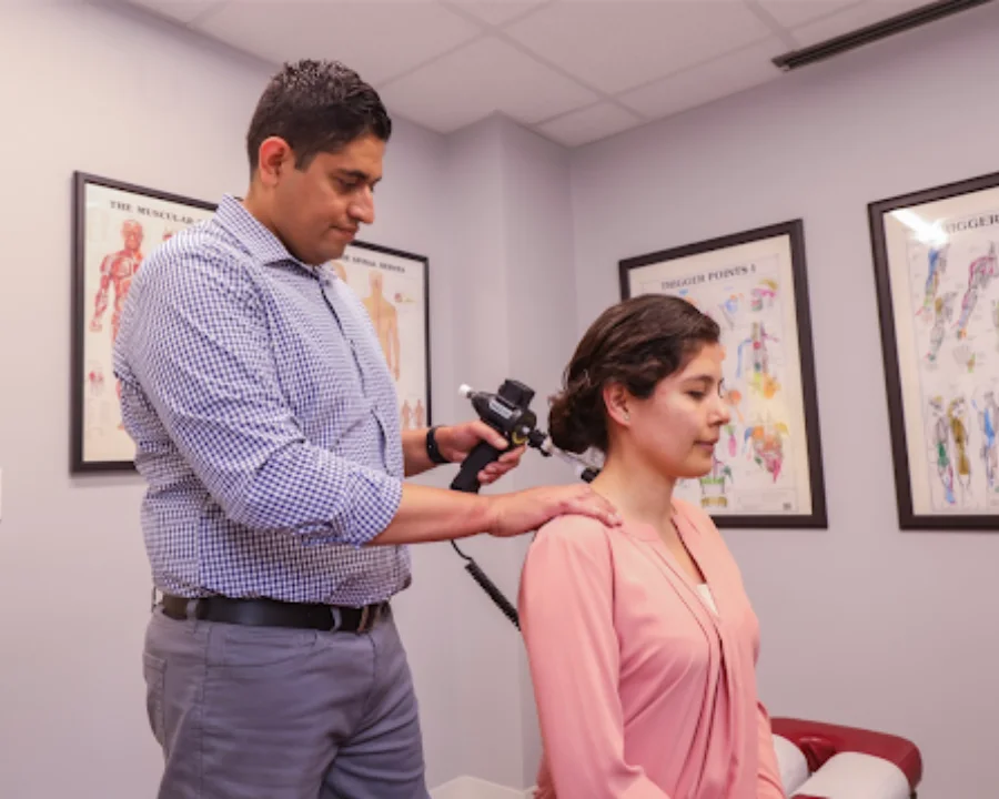 Chiropractor using a handheld instrument to perform an adjustment on a seated female patient's neck in a chiropractic office