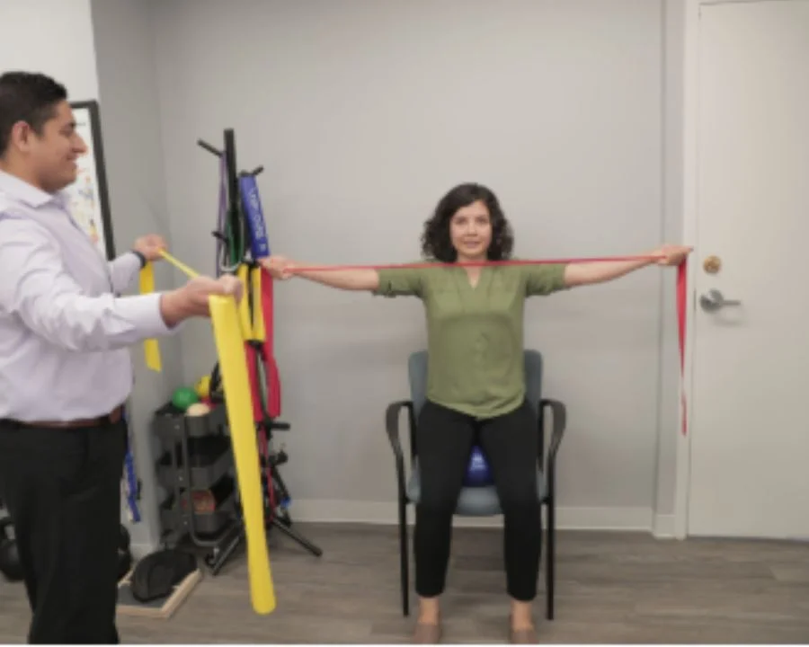 Chiropractor assisting a seated patient with resistance band shoulder exercises during a physical therapy session